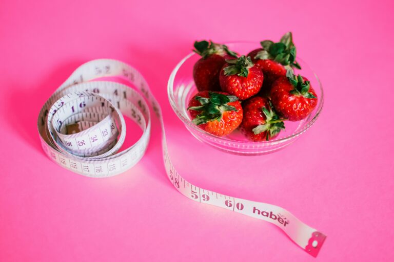 Ruban à mesurer enroulé blanc et bol de fraises rouges fraîches sur fond rose, illustrant les bienfaits jeûne intermittent : perte de poids rapide, réduction graisse abdominale, stabilisation glycémie, autophagie cellulaire, énergie boostée et santé cardiovasculaire améliorée