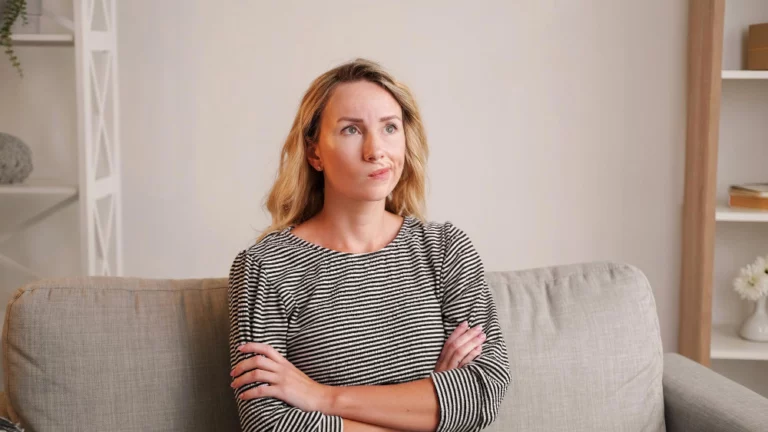 Femme d'une cinquantaine d'années assise sur un canapé, bras croisés et regard pensif, illustrant la fatigue hormonale et le manque d'énergie liés à la ménopause