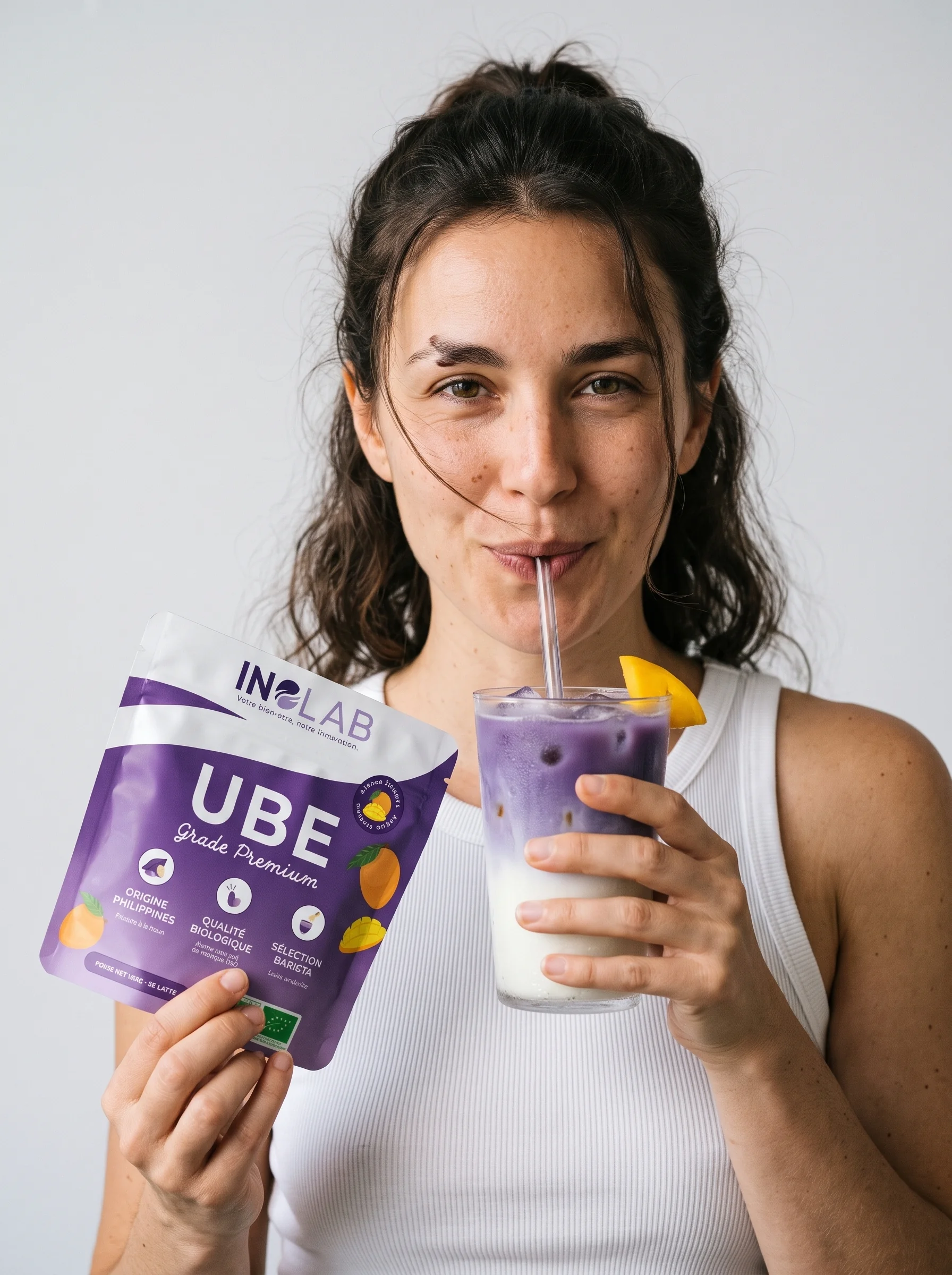 Femme riante assise en tailleur avec un pochon d'Ube Mangue Inolab et un verre d'ube latte
