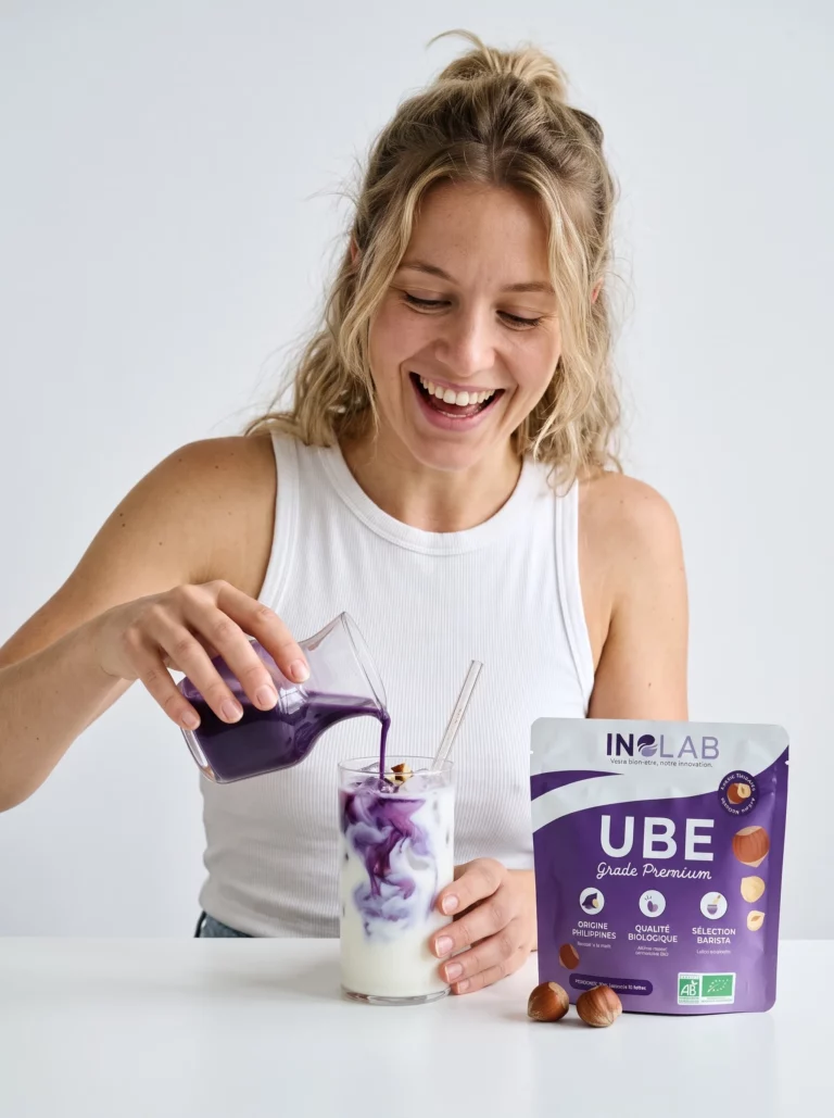 Femme préparant un ube latte à la noisette en versant du lait d'ube violet, pochon d'Ube Noisette Inolab à côté
