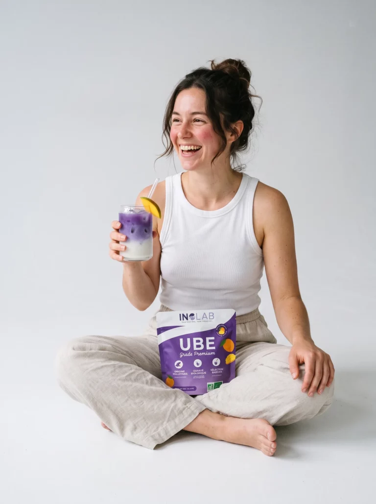 Femme riante assise en tailleur avec un pochon d'Ube Mangue Inolab et un verre d'ube latte